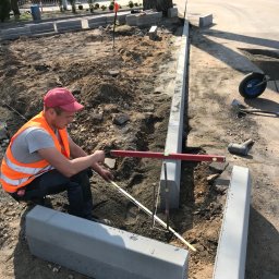 PIXEL BUD VOLODYMYR ZATSERKOVNYI SP.K. - Pracownik w kamizelce odblaskowej i czerwonej czapce mierzy poziomnicą i miarką rozmieszczenie betonowych obrzeży chodnika na gruncie, przygotowując się do ich osadzenia. W tle widoczna budowa...