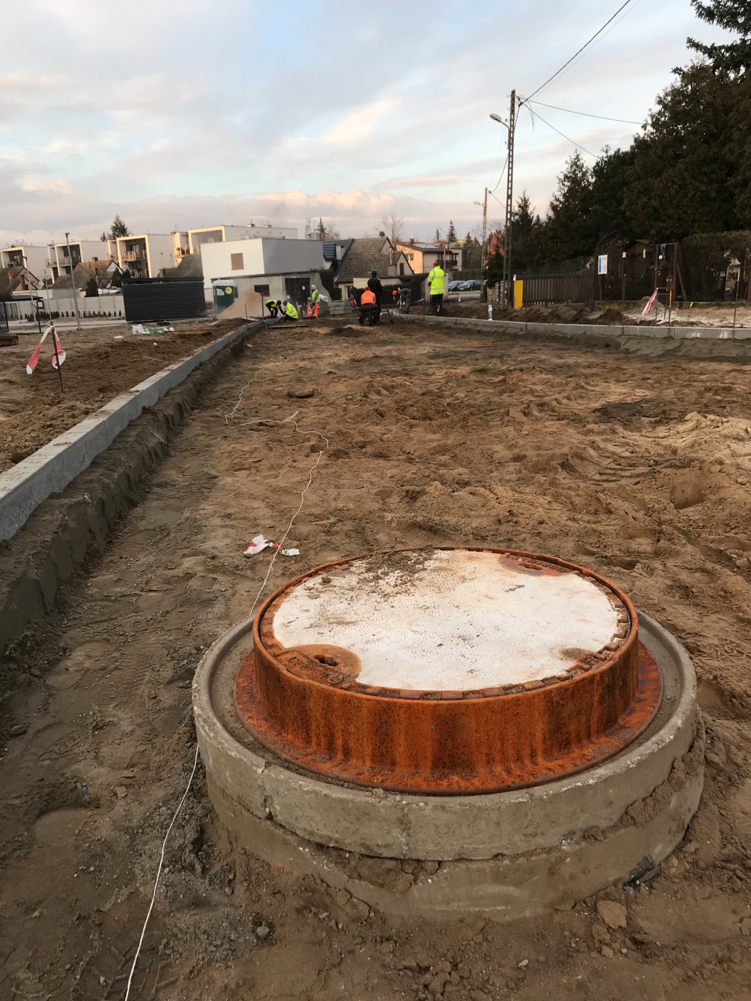 Przygotowanie terenu pod budowę chodnika z widocznym włazem kanalizacyjnym z pomarańczowym pierścieniem i pracownikami w tle.