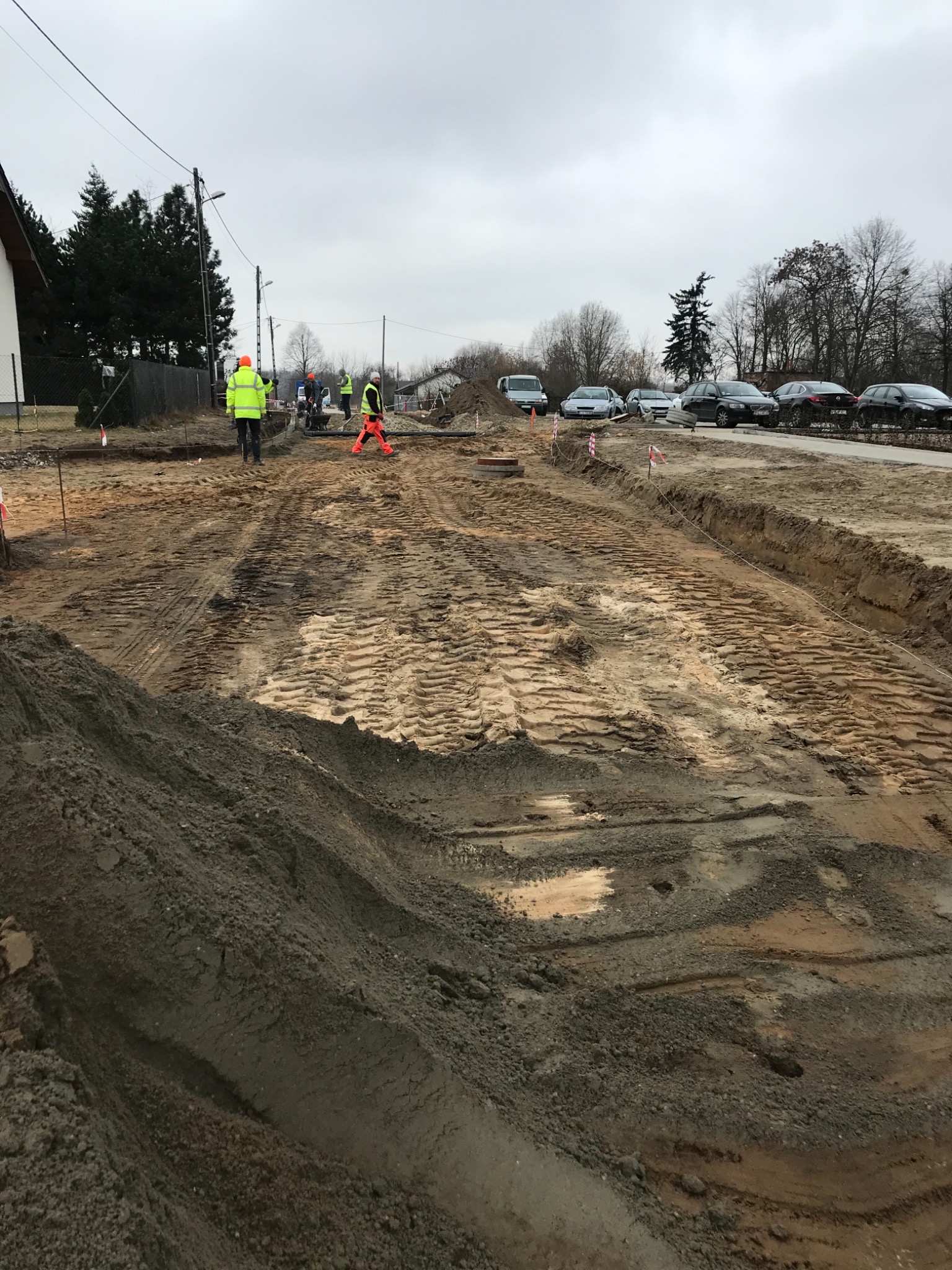 Przygotowanie terenu pod budowę drogi: wyrównany teren z widocznymi śladami opon, pracownicy w odblaskowych ubraniach, samochody w tle, pochmurne niebo.
