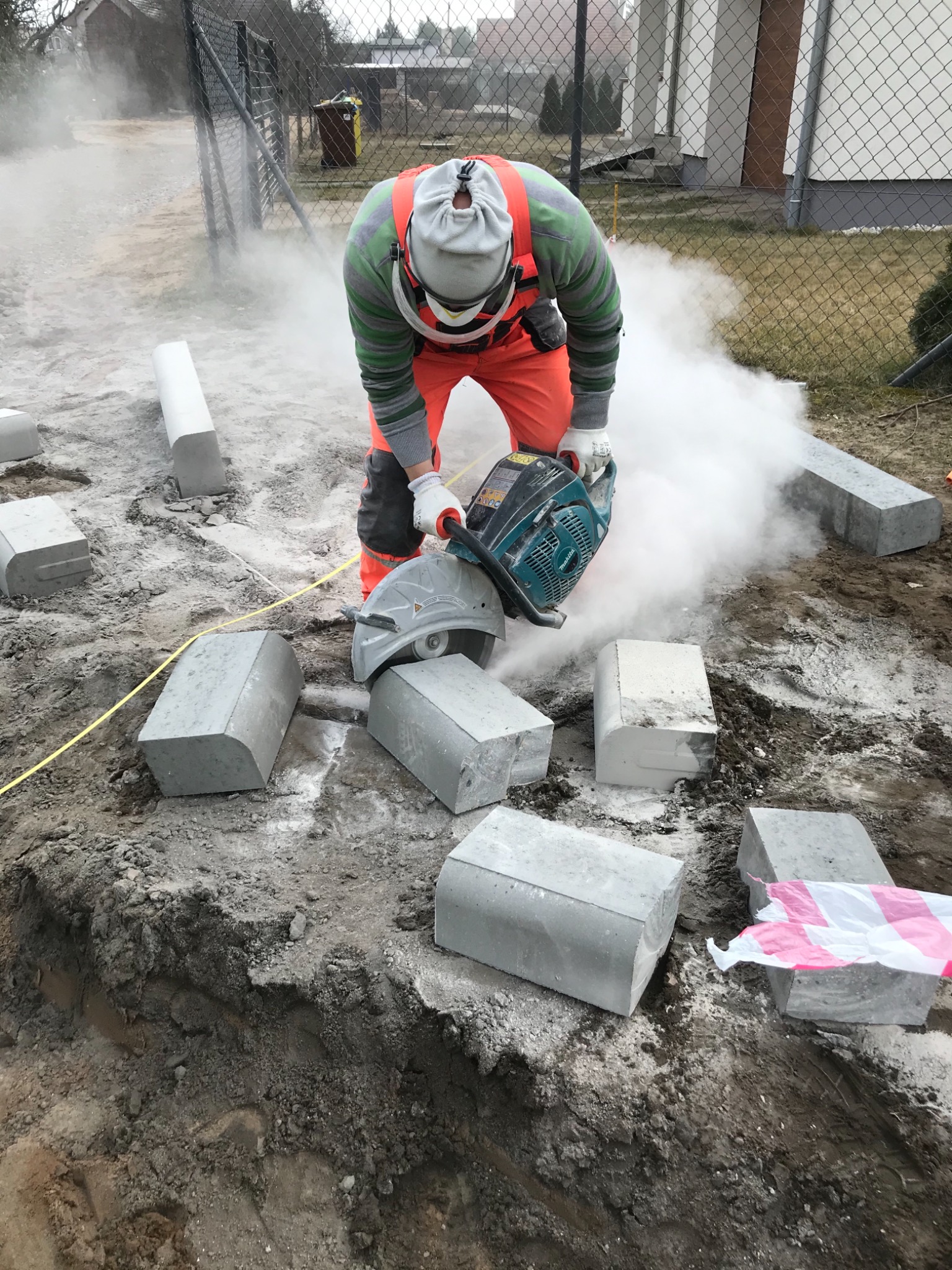 Pracownik w pomarańczowym kombinezonie tnie szlifierką kątową betonowy obrzeżnik, unosząc chmurę pyłu, widoczne inne obrzeżniki i fragment wykopu.