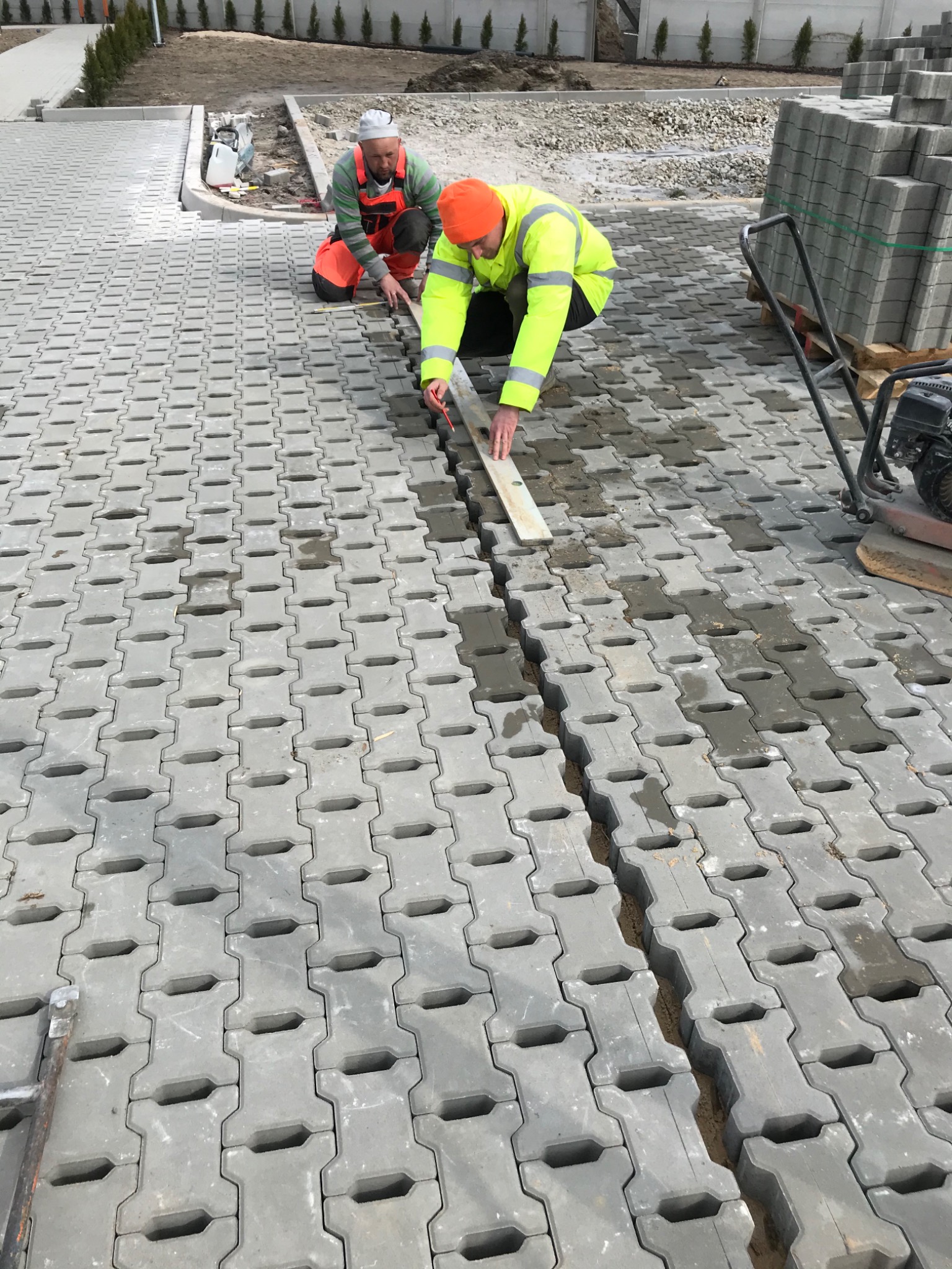 Układanie szarej kostki brukowej typu I na podsypce piaskowej, dwóch pracowników wyrównuje poziom za pomocą poziomicy i łaty, w tle paleta z kostką i ubijarka wibracyjna.