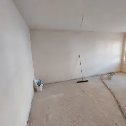 Pokój po tynkowaniu maszynowym, widoczna gładka, jasna ściana, wiadro z resztkami materiałów budowlanych, rura i miotła oparta o ścianę.