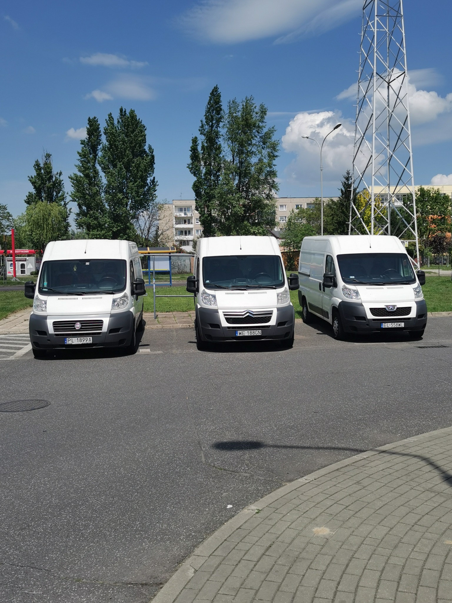 Trzy białe busy różnych marek (Fiat, Citroen, Peugeot) zaparkowane obok siebie na parkingu w słoneczny dzień. Widoczny budynek w tle i wieża telekomunikacyjna.