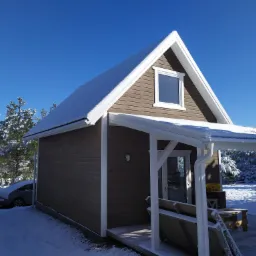 Niewielki, brązowy dom z białymi elementami wykończeniowymi, pokryty śniegiem, na tle ośnieżonego lasu i błękitnego nieba.