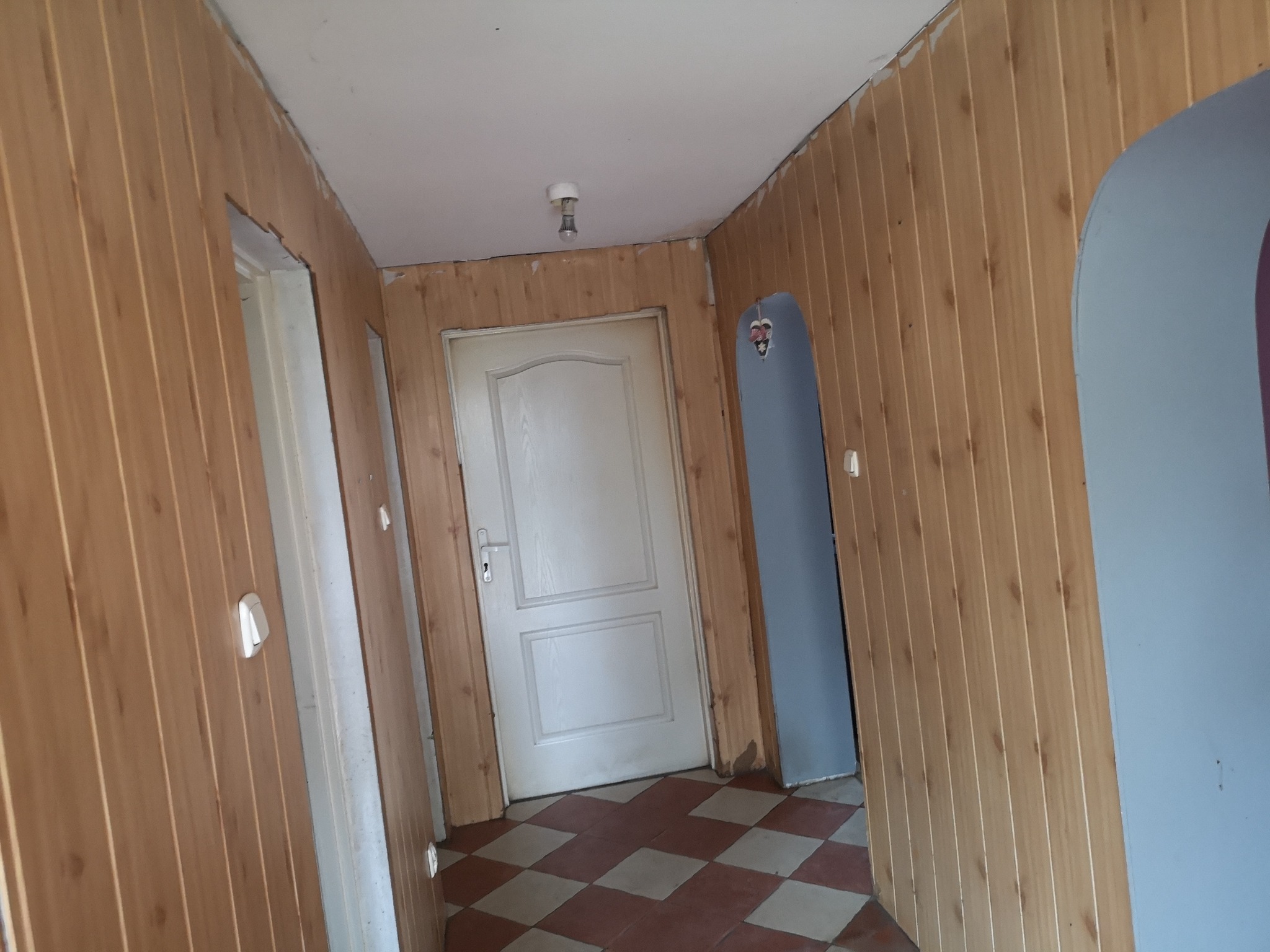 Korytarz wykończony boazerią imitującą drewno, z białymi drzwiami i szachownicą z płytek na podłodze, przed renowacją.