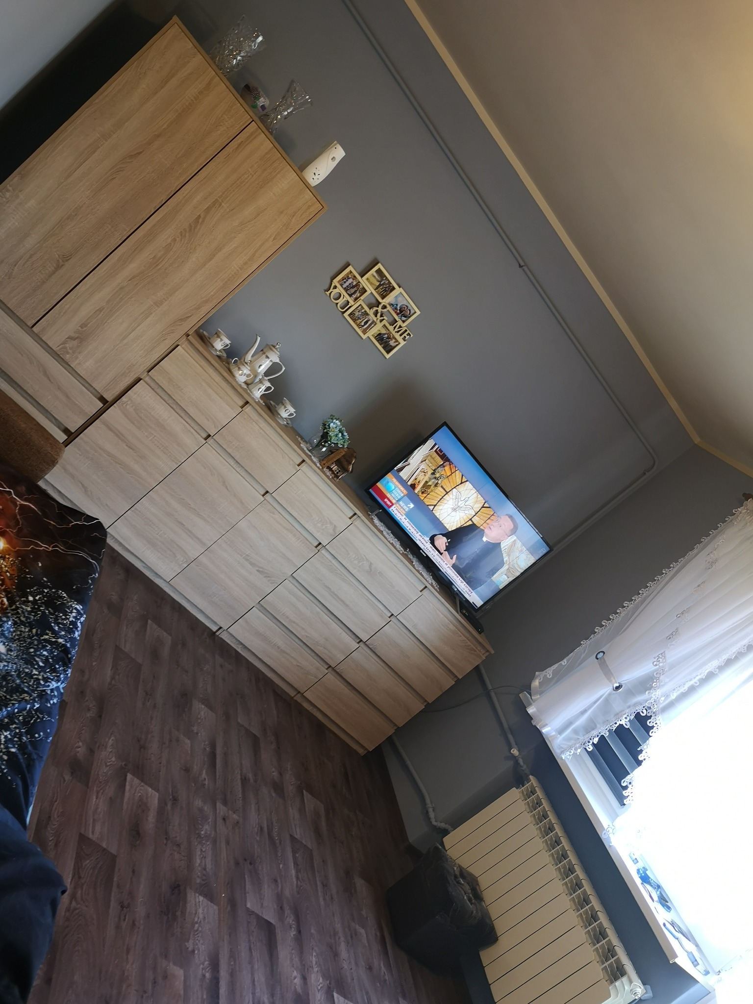 Komoda z szufladami w jasnym odcieniu drewna, z telewizorem na blacie, ustawiona w pokoju z szarą ścianą i drewnianą podłogą.