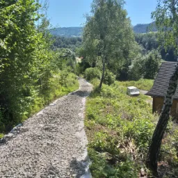 Droga gruntowa wysypana szarym żwirem, zabezpieczona geowłókniną, prowadząca przez teren zalesiony w kierunku zabudowań i zaparkowanego białego samochodu dostawczego w oddali, widok z góry.