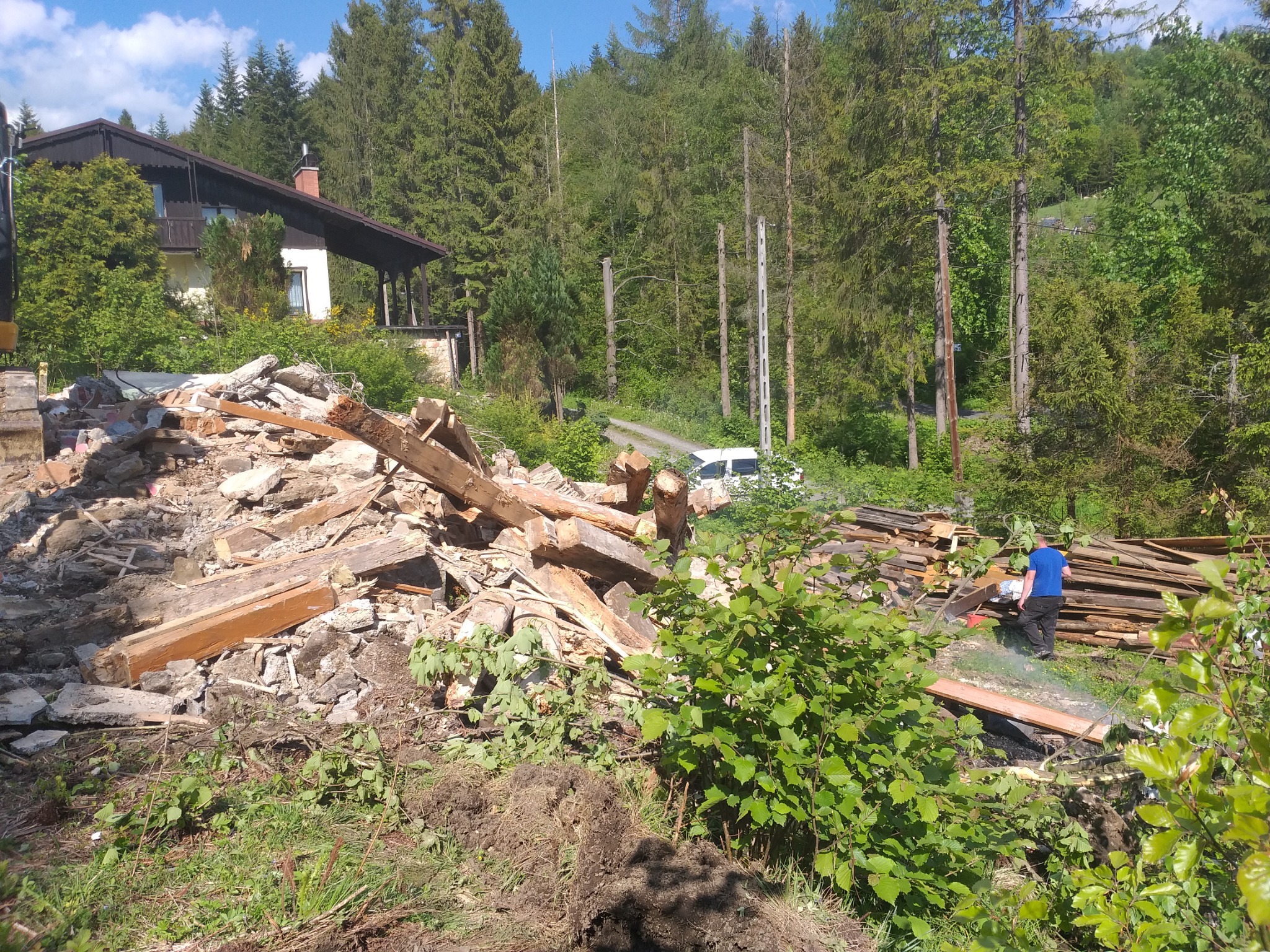 Góra gruzu i drewnianych belek po rozbiórce budynku, zalesione wzgórze w tle, pracownik w niebieskiej koszulce porządkujący drewno.