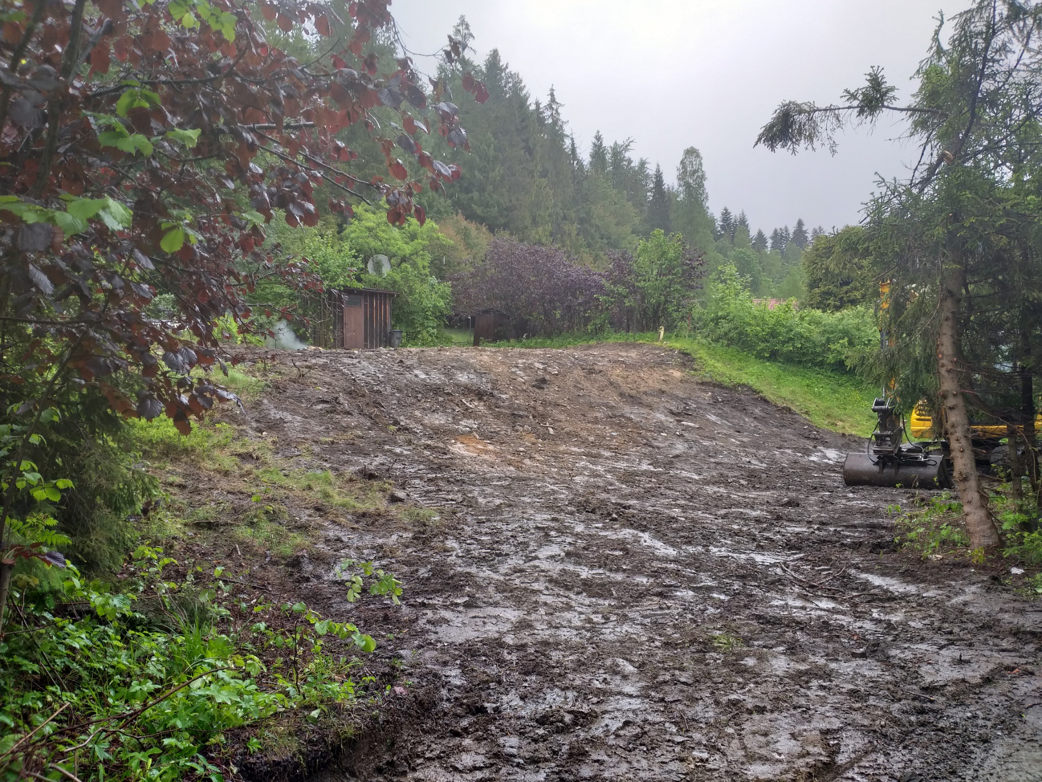 Teren po wyrównaniu w Istebnej, z widocznymi śladami opon i małą koparką w tle, otoczony drzewami i krzewami w pochmurny dzień.