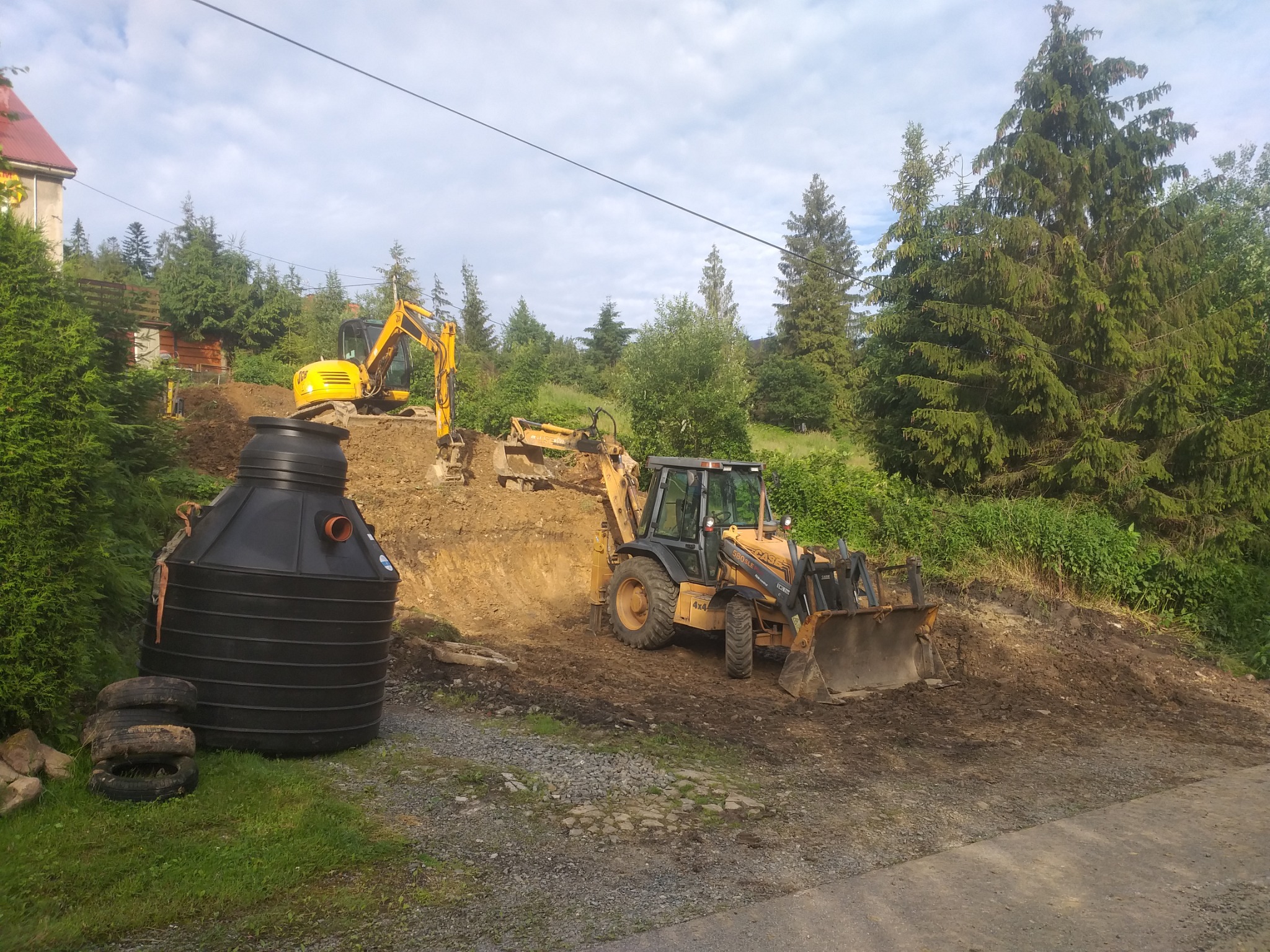 Dwa żółte i czarne traktory na placu budowy w pagórkowatym terenie, jeden z czarnym zbiornikiem na pierwszym planie i stertą opon obok, drzewa w tle.