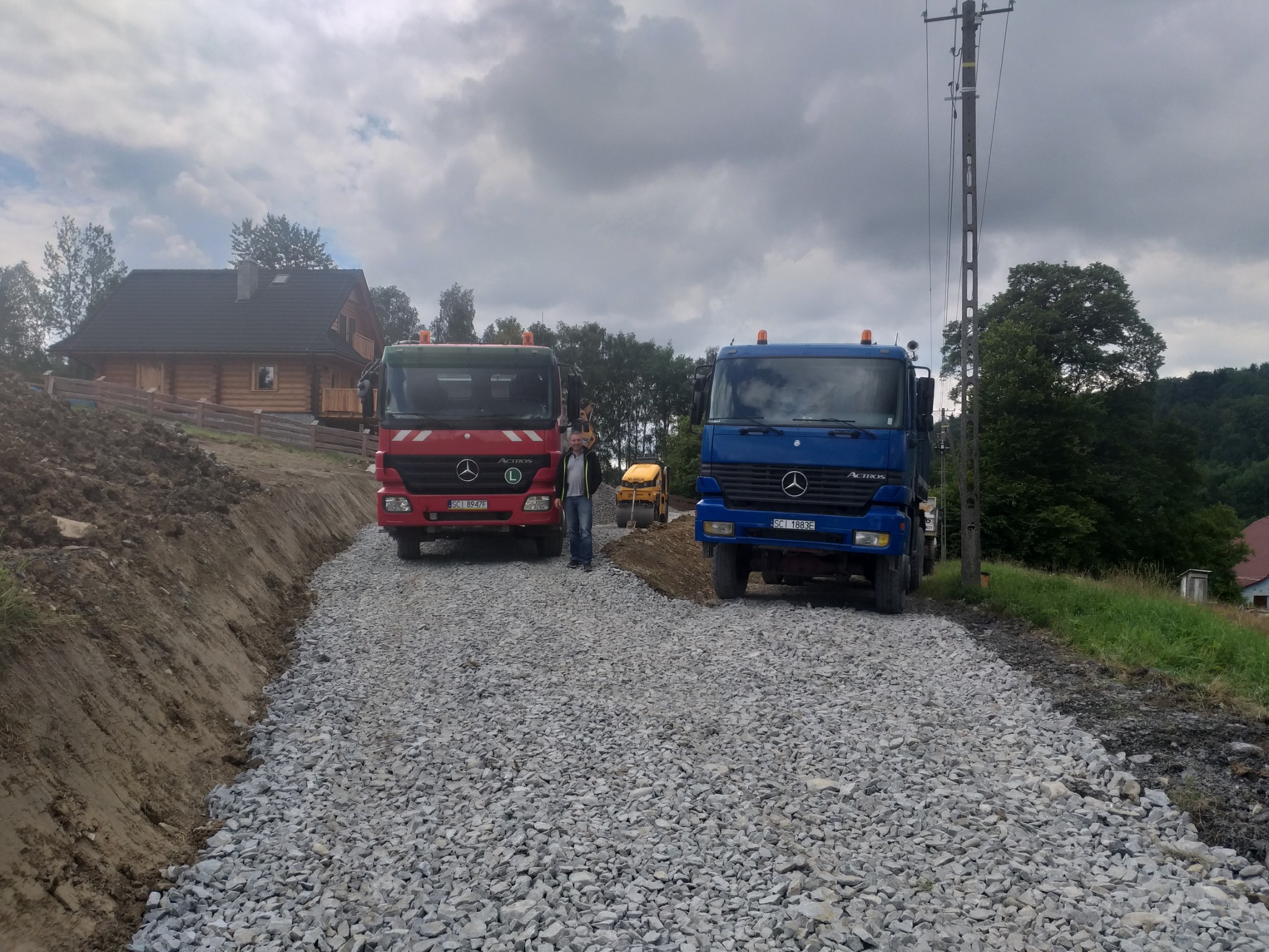 Dwa samochody ciężarowe (czerwony i niebieski) stoją na nowo utwardzonej drodze żwirowej, w tle widać mały walec drogowy i drewniany dom. Na pierwszym planie mężczyzna w dżinsach.