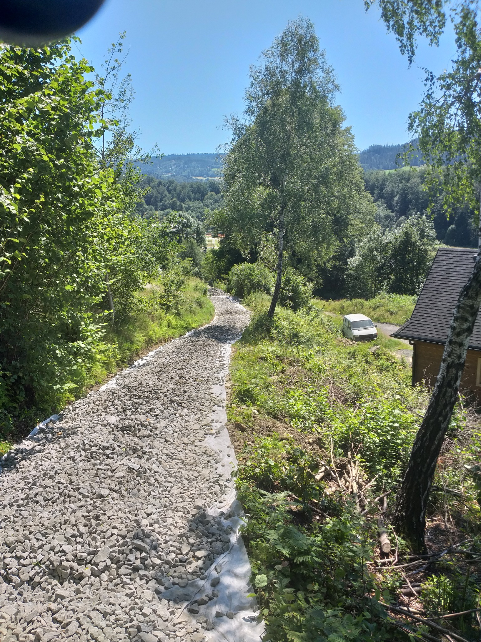 Droga gruntowa wysypana szarym żwirem, zabezpieczona geowłókniną, prowadząca przez teren zalesiony w kierunku zabudowań i zaparkowanego białego samochodu dostawczego w oddali, widok z góry.