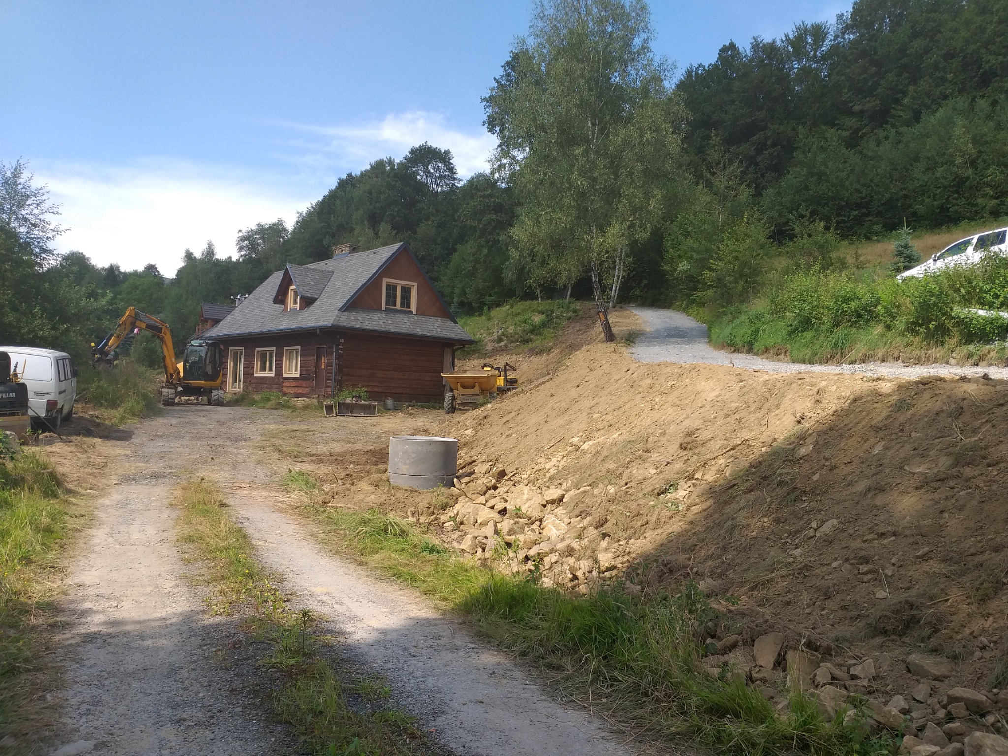 Żółta koparka Caterpillar przy drewnianym domu, przygotowanie terenu pod budowę, widoczny wykop, hałda ziemi i betonowy krąg.