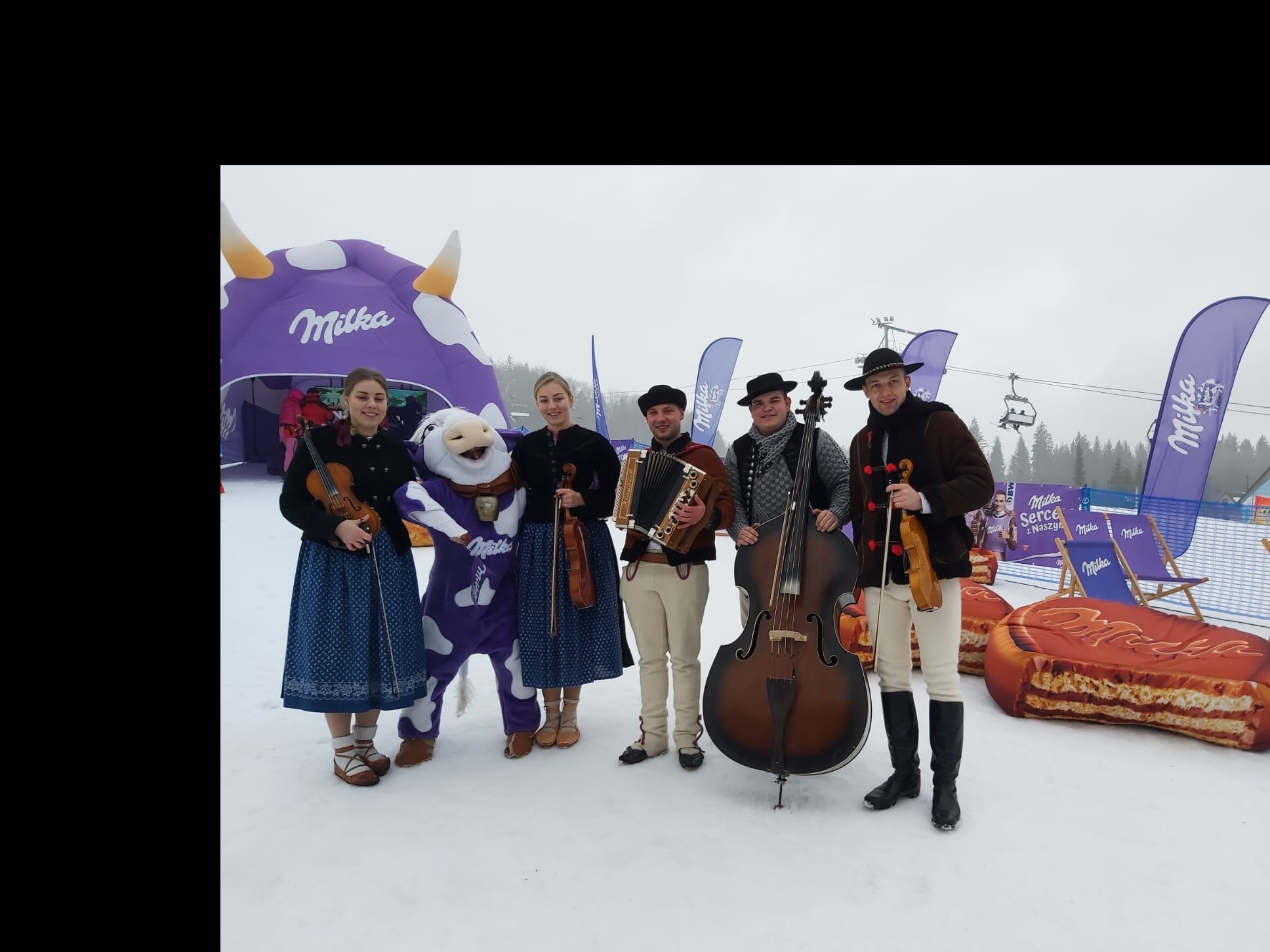 Grupa górali w tradycyjnych strojach, z maskotką Milki i instrumentami muzycznymi (skrzypce, akordeon, kontrabas) na tle zimowego krajobrazu i dmuchanych rekwizytów Milki.