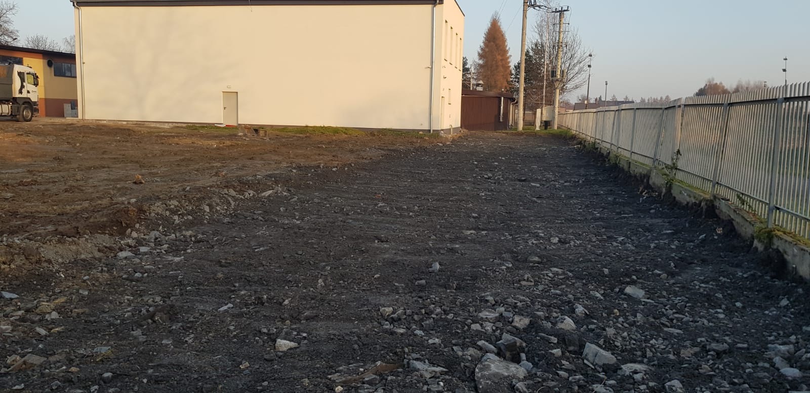 Przygotowany teren pod budowę z widoczną wyrównaną ziemią, kamieniami i budynkiem w tle, ogrodzenie po prawej stronie.