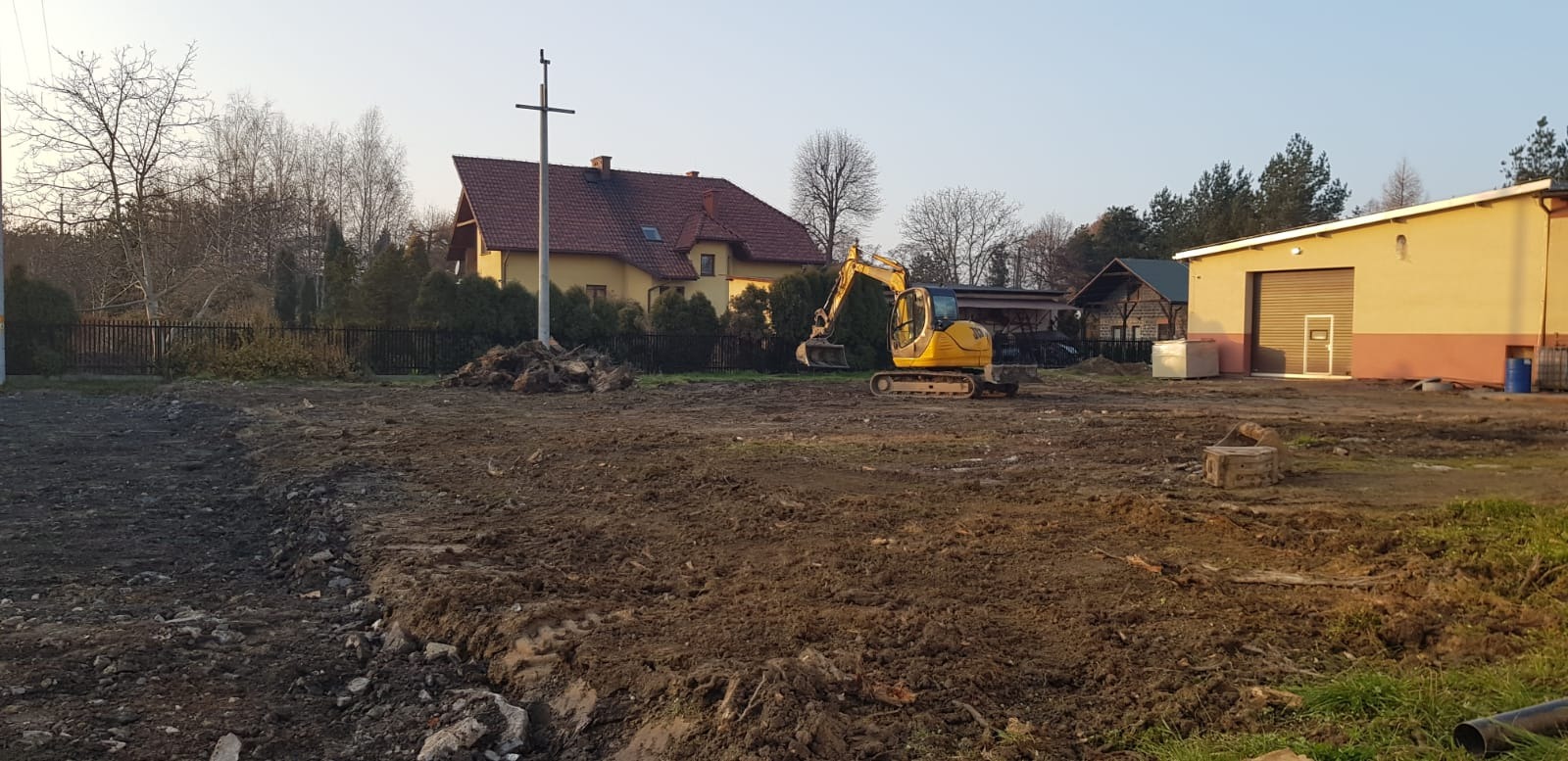 Żółta koparka na placu budowy wyrównuje teren, w tle dom z czerwonym dachem i budynek gospodarczy z bramą rolowaną.