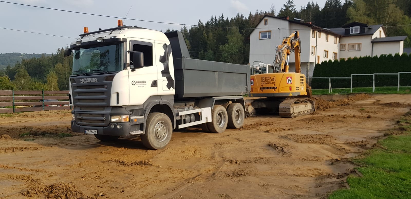 Ciężarówka Scania z naczepą obok żółtej koparki Liebherr 924 na placu budowy z wykopaną ziemią, w tle budynek mieszkalny i zalesione wzgórza.