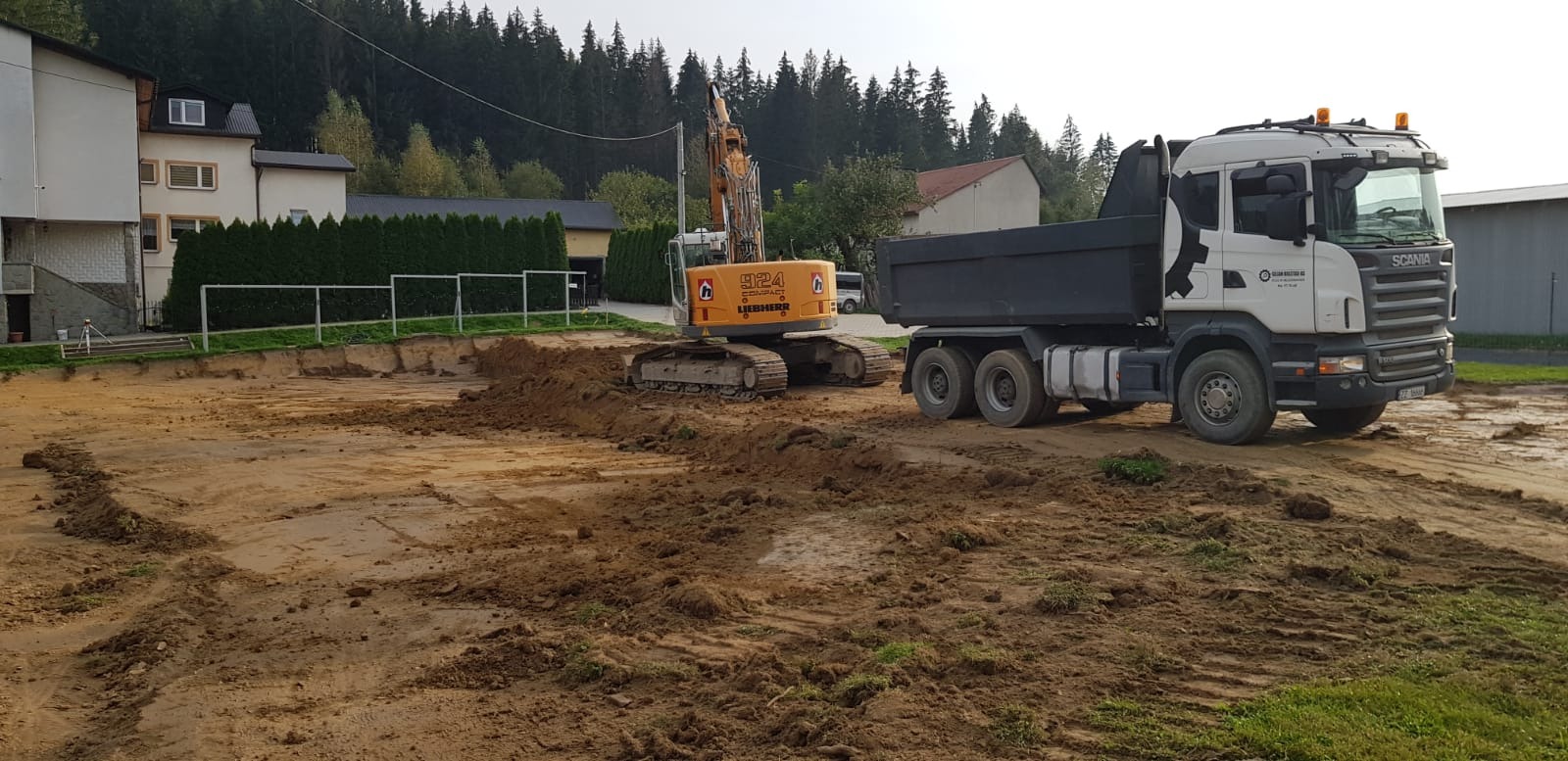 Żółta koparka Liebherr 924 i biała ciężarówka Scania na placu budowy, wykopana ziemia, w tle budynki i drzewa.