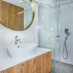 Nowoczesna łazienka z owalną umywalką nablatową, lustrem w złotej ramie, kabiną prysznicową ze szklanymi drzwiami i marmurowymi płytkami na ścianach.