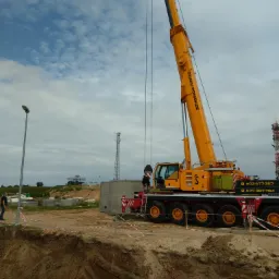 Żółty dźwig samochodowy firmy Dźwigi Pomorze opuszcza prefabrykowany betonowy element do wykopu w ziemi, w tle widoczna wieża telekomunikacyjna i pochmurne niebo.