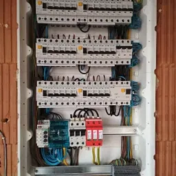 Rozdzielnica elektryczna w trakcie montażu, widoczne zabezpieczenia nadprądowe, ograniczniki przepięć, listwy zaciskowe, przewody w kolorach żółto-zielonym, niebieskim i czarnym, ściana z czerwonej...