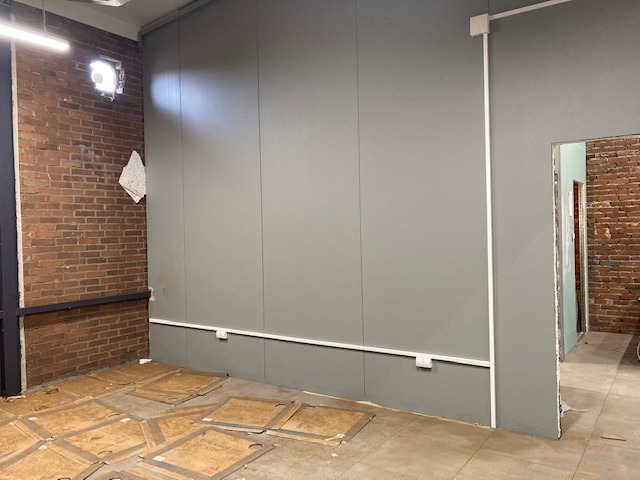 Wnętrze w trakcie remontu: szara ściana z listwami elektrycznymi, odsłonięta cegła, podłoga z kartonami ochronnymi, widoczne gniazdka elektryczne.