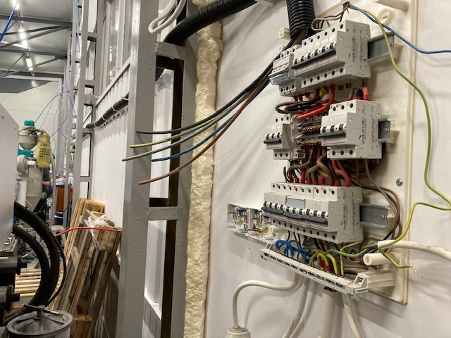 Szafa elektryczna z rozłączonymi przewodami w trakcie montażu w hali przemysłowej w Warszawie, widoczne zabezpieczenia nadprądowe i kable różnych kolorów.