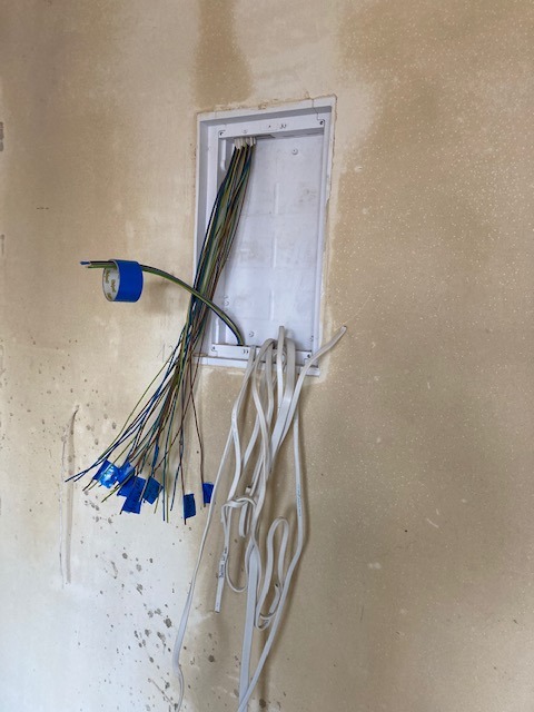 Ściana w trakcie remontu z widoczną puszką instalacyjną, z której wychodzą przewody elektryczne, zabezpieczone niebieską taśmą izolacyjną.