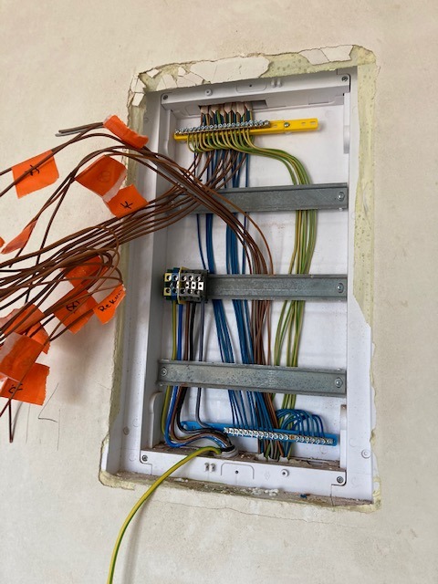 Otwarta skrzynka elektryczna w ścianie z widocznymi przewodami oznaczonymi pomarańczowymi etykietami, listwy zaciskowe i metalowe szyny montażowe, ściana w trakcie remontu.