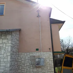 Zewnętrzna skrzynka elektryczna z otwartymi drzwiczkami, zamontowana na elewacji budynku z dekoracyjną okładziną kamienną, widoczna rura instalacyjna i fragment samochodu dostawczego.