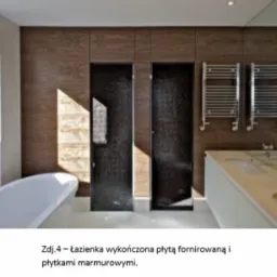 Nowoczesna łazienka z wanną wolnostojącą, ścianą wykończoną płytami fornirowanymi w odcieniach brązu, dwoma czarnymi, mozaikowymi kabinami prysznicowymi i białym blatem z umywalką.