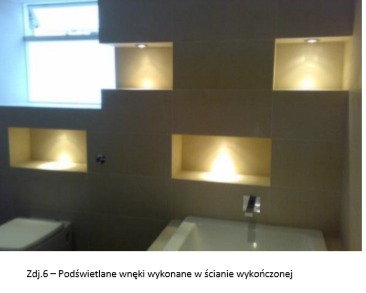 Beżowa ściana w łazience z wnękami oświetlonymi punktowo, widoczna toaleta i fragment wanny, nowoczesny design.