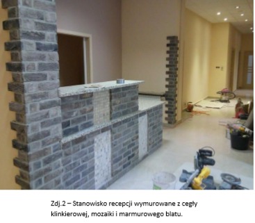 Stanowisko recepcji wykończone cegłą klinkierową, mozaiką i blatem marmurowym. Widoczny fragment ściany z cegły oraz podłoga w trakcie prac wykończeniowych.