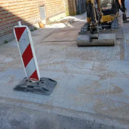 Mini koparka na placu budowy w Bielsku-Białej, przygotowuje teren pod nową nawierzchnię z kostki brukowej, widoczny słupek ostrzegawczy i rozsypany piasek.