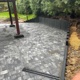 Ułożona szara kostka brukowa na posesji z odpływem liniowym, obrzeżami i niskim murem oporowym w tle, widoczne drzewa i zieleń.