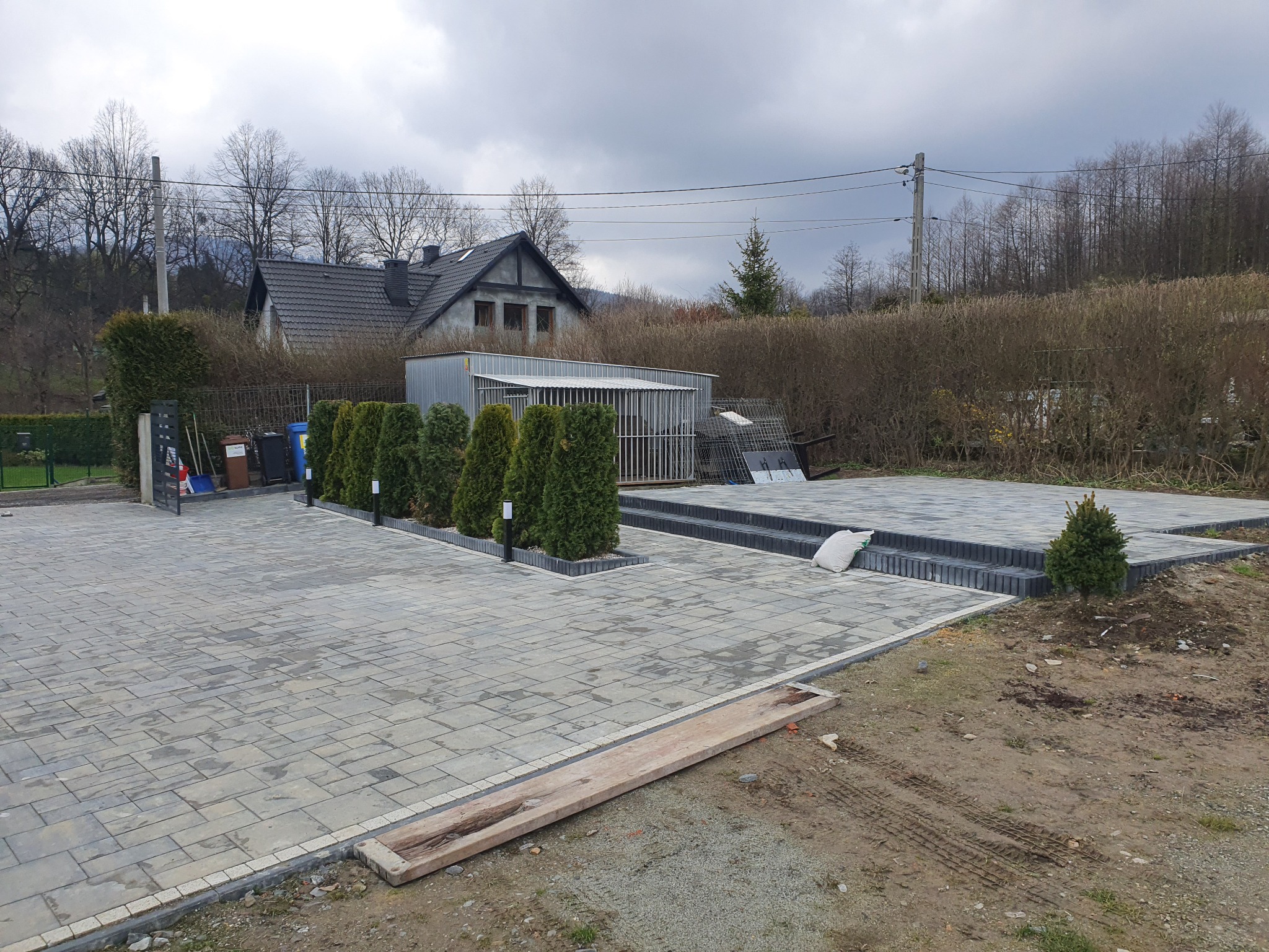 Ukończona nawierzchnia z kostki brukowej w Bielsku-Białej, z obrzeżami i schodami z tego samego materiału, uzupełniona tujami i oświetleniem, na tle domu i ogrodu.