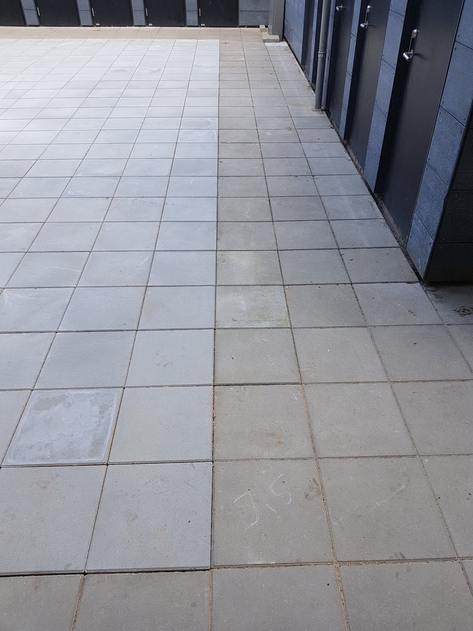 Szara kostka brukowa ułożona w kwadraty, widoczne ślady po kredzie na kilku elementach, obok ściana z ciemnymi drzwiami.