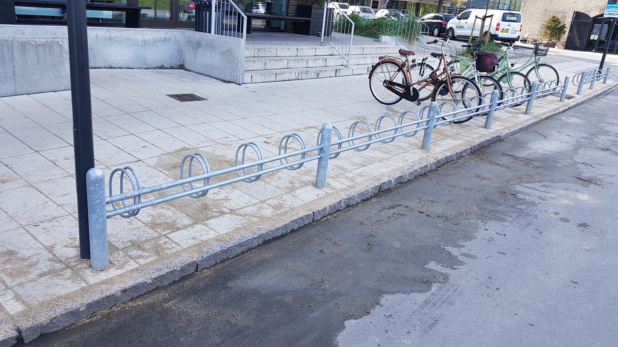 Szary, metalowy stojak rowerowy z zaparkowanymi rowerami na tle chodnika wyłożonego kostką. W tle budynek i zaparkowane samochody.