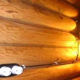 Dwa gniazdka elektryczne natynkowe z podwójnym otworem i kablem w oplocie, zamontowane na ścianie z drewnianych bali, obok dekoracyjnej lampy ściennej z dwoma kloszami.