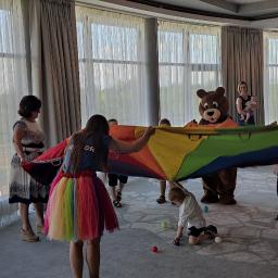 Holiday organizacja imprez - Animatorka w tęczowej spódniczce i pluszowy miś trzymają kolorową chustę animacyjną podczas zabawy z dziećmi w przestronnym pomieszczeniu z dużymi oknami.