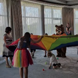 Animatorka w tęczowej spódniczce i pluszowy miś trzymają kolorową chustę animacyjną podczas zabawy z dziećmi w przestronnym pomieszczeniu z dużymi oknami.