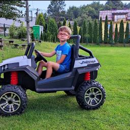 Holiday organizacja imprez - Chłopiec w okularach siedzi za kierownicą białego elektrycznego pojazdu terenowego na zielonym trawniku przed budynkami.