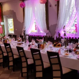 Uroczysty stół w Borku Starym, nakryty białym obrusem, z dekoracjami kwiatowymi, szklanymi naczyniami i różowymi akcentami, przygotowany do przyjęcia gości w sali z czerwonymi wzorzystymi ścianami...