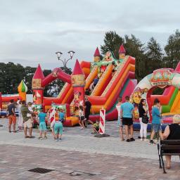 Holiday organizacja imprez - Dwa duże, dmuchane place zabaw z motywami z bajek, otoczone grupą dzieci i dorosłych na placu z kostki brukowej.