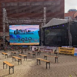 Przygotowania do plenerowej imprezy: scena z telebimem wyświetlającym nazwę wydarzenia 'Dzień Bieszczadzki 2020', rzędy drewnianych ławek dla publiczności, białe namioty w tle, budynki w tle.
