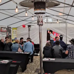 Wnętrze białego namiotu eventowego z metalowym ogrzewaczem, podwieszonymi żarówkami i stolikami, przy których siedzą goście, w tle widoczne roll-upy reklamowe różnych firm.