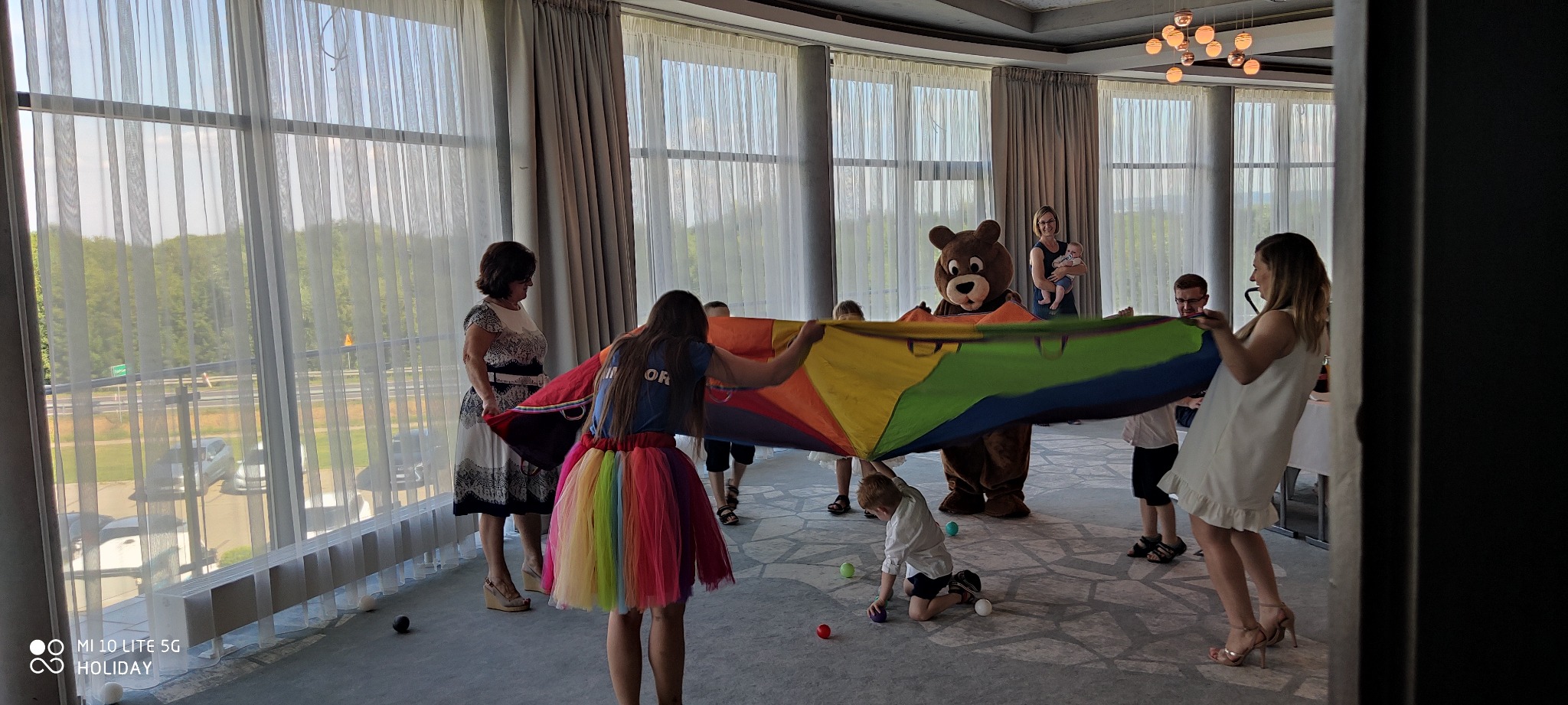 Animatorka w tęczowej spódniczce i pluszowy miś trzymają kolorową chustę animacyjną podczas zabawy z dziećmi w przestronnym pomieszczeniu z dużymi oknami.