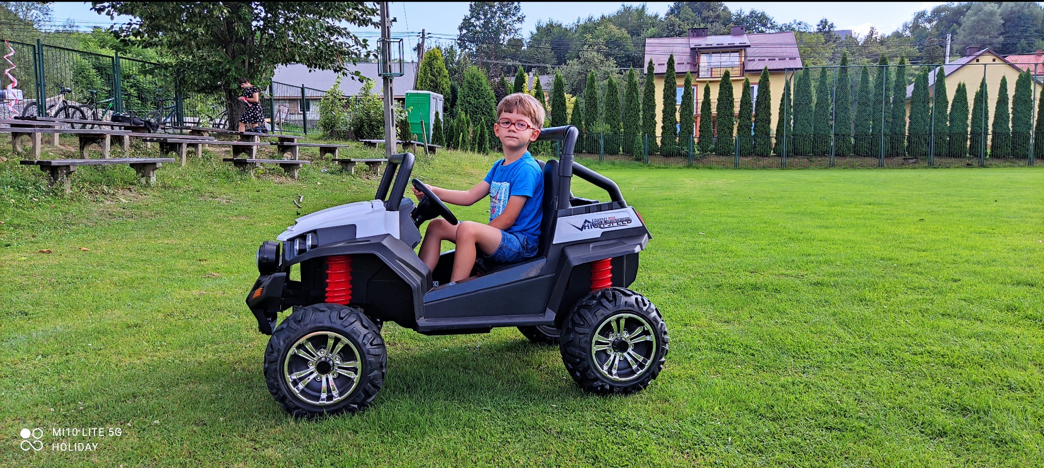 Chłopiec w okularach siedzi za kierownicą białego elektrycznego pojazdu terenowego na zielonym trawniku przed budynkami.