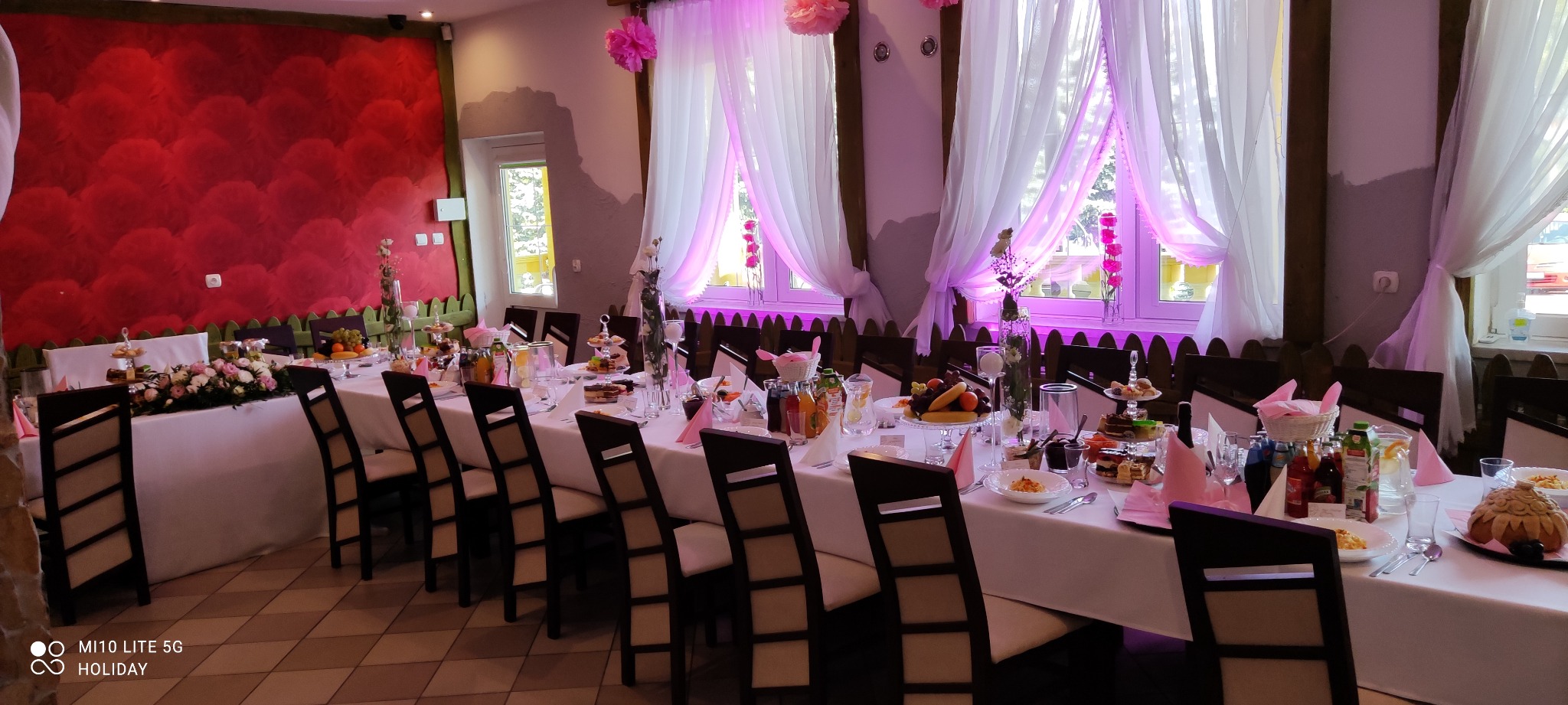 Uroczysty stół w Borku Starym, nakryty białym obrusem, z dekoracjami kwiatowymi, szklanymi naczyniami i różowymi akcentami, przygotowany do przyjęcia gości w sali z czerwonymi wzorzystymi ścianami...