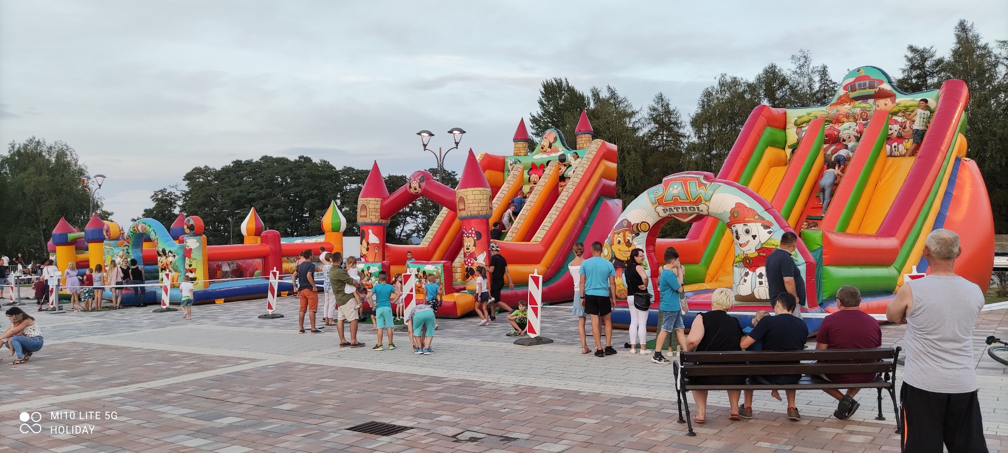 Dwa duże, dmuchane place zabaw z motywami z bajek, otoczone grupą dzieci i dorosłych na placu z kostki brukowej.